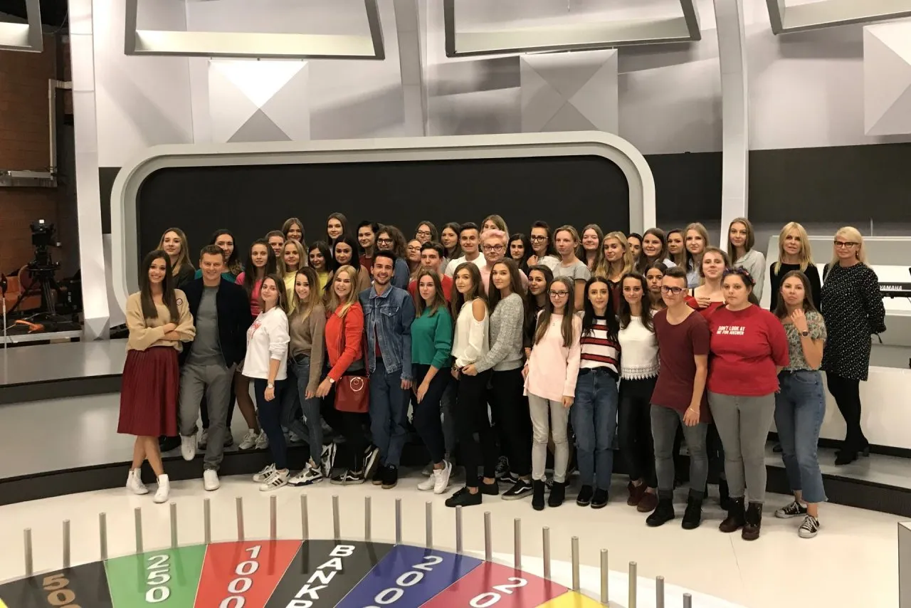 Grupa ludzi pozuje do zdjęcia w studiu telewizyjnym, gdzie znajduje się koło fortuny.