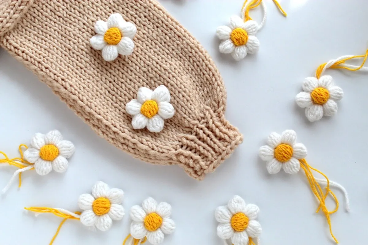 Urocze dekoracje na szydełku w kształcie stokrotek zdobią beżowy, dzianinowy rękaw.
