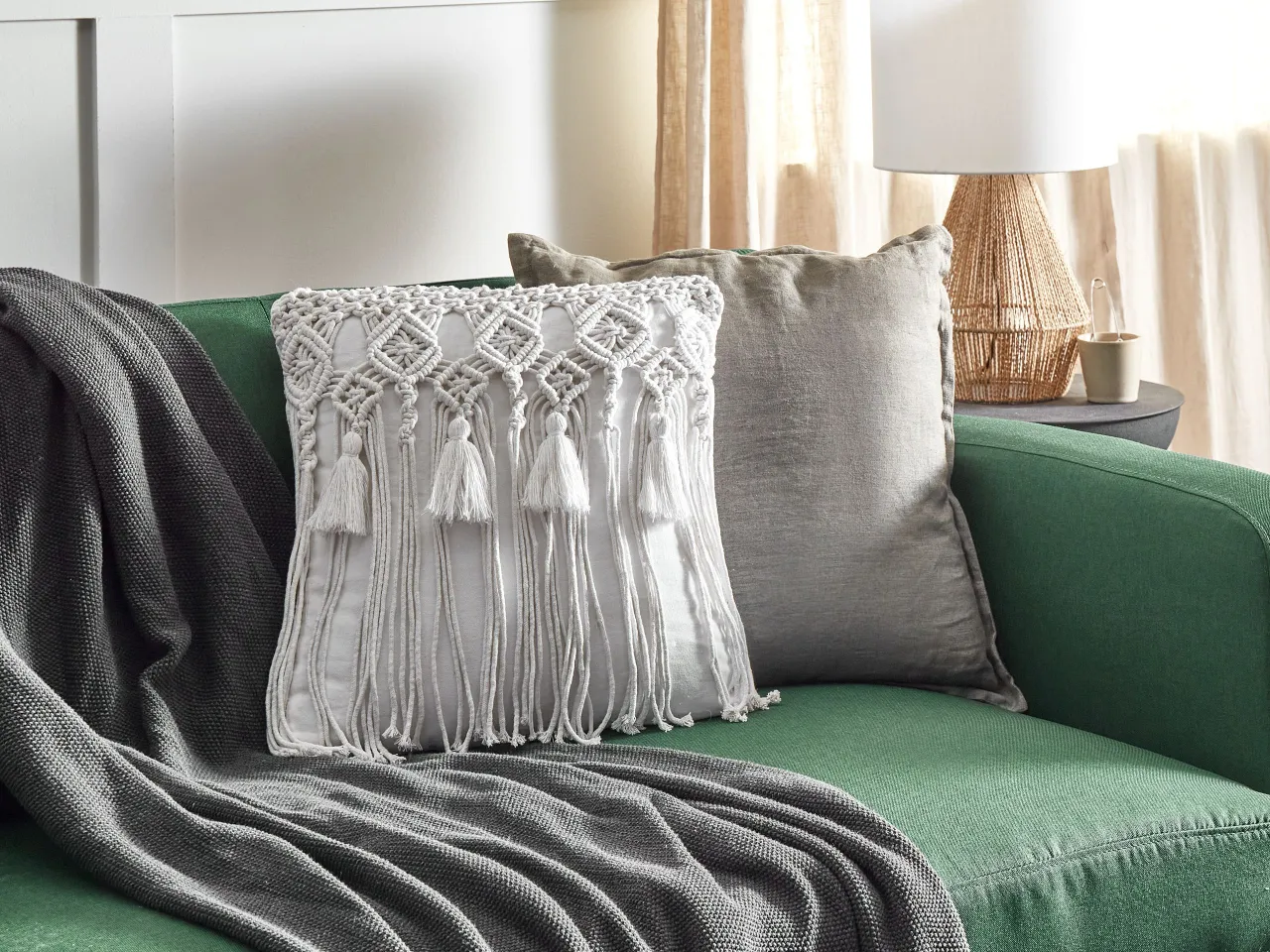 Biała poduszka makrama krok po kroku, ozdobiona frędzlami, leży na zielonej kanapie obok szarego koca i drugiej poduszki.