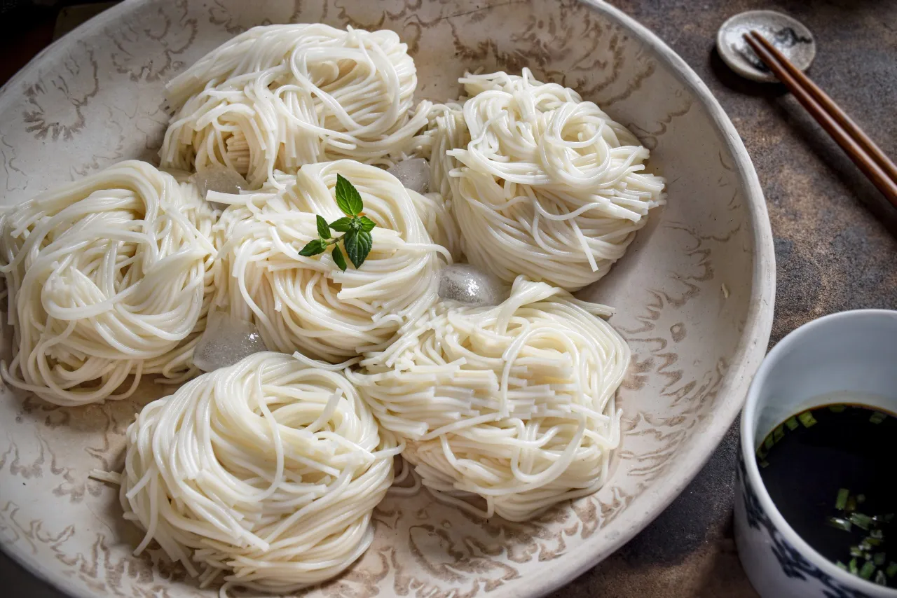 Schłodzone kluski somen w misce, gotowe do zanurzenia w sosie do tempury. Na talerzyku pałeczki i miseczka z ciemnym sosem.