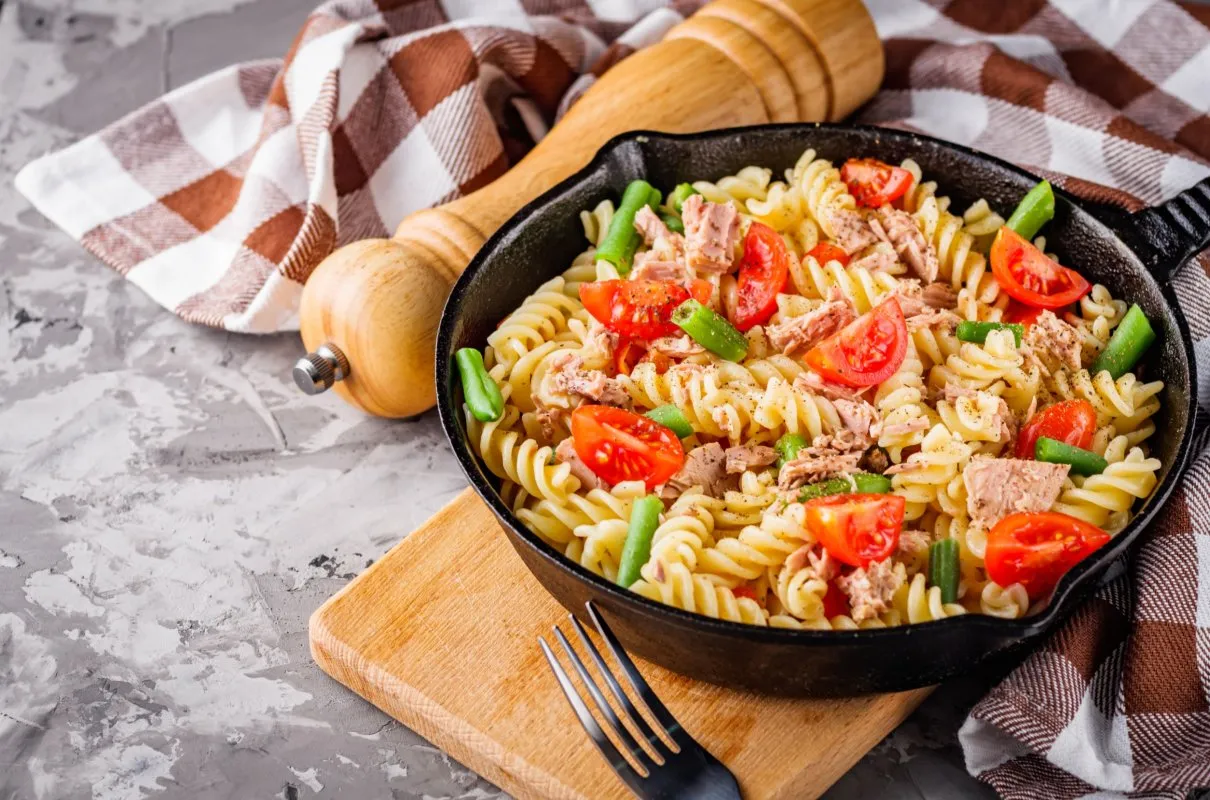 Makaron z tuńczykiem i pomidorkami, danie, co każdy potrafi ugotować. Podane w patelni, obok młynek do pieprzu i kratkowana ściereczka.