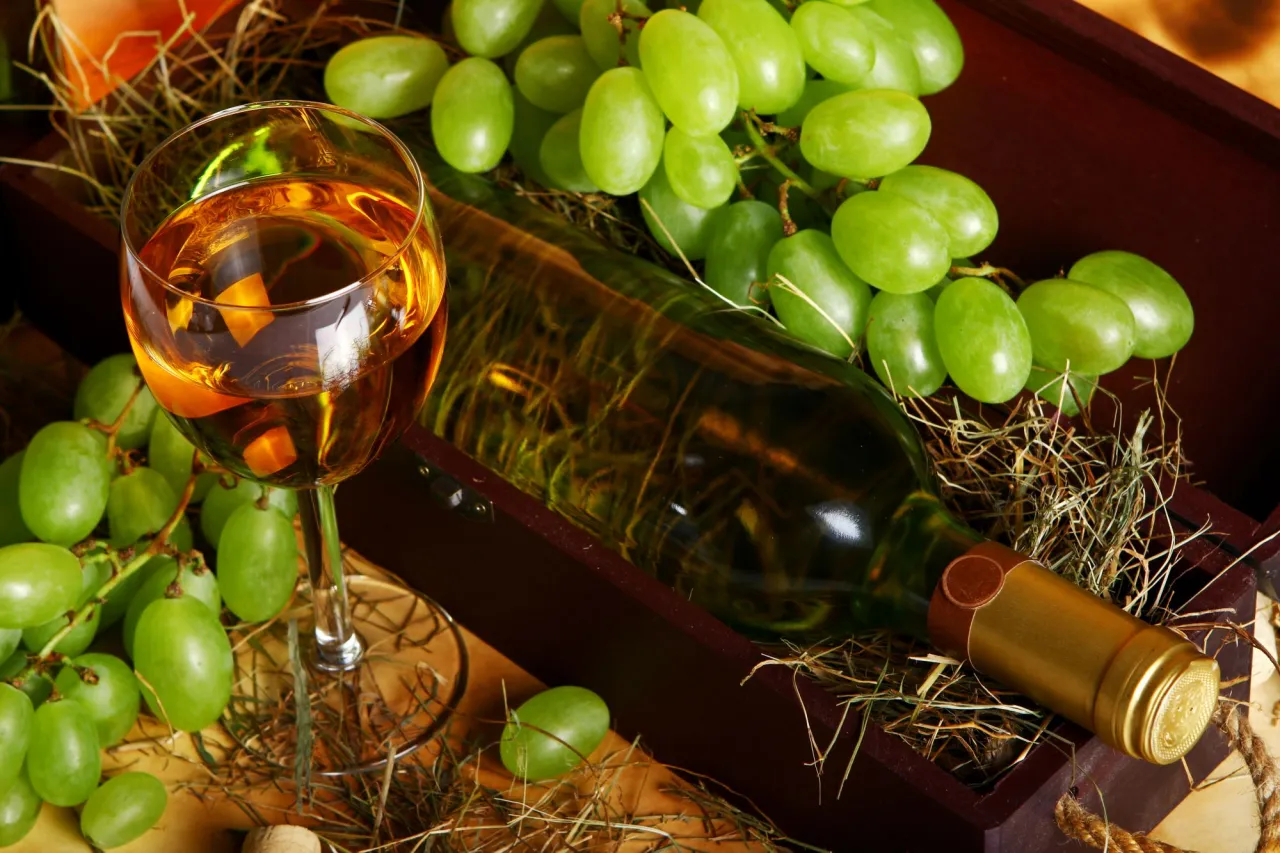 W kieliszku złocisty trunek, obok butelka i winogrona. Zastanawiasz się, czemu wino nie jest wegańskie? Proces klarowania może wykorzystywać produkty zwierzęce.