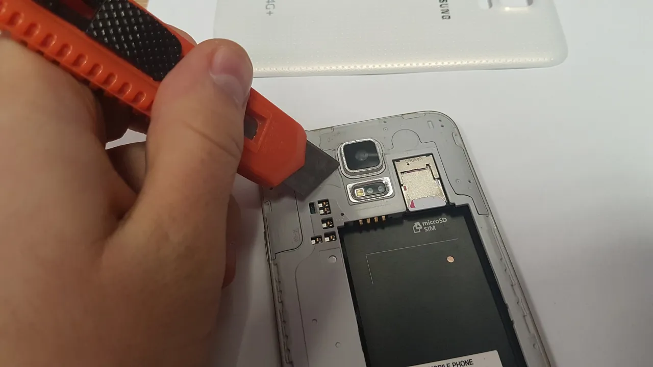 Dłoń z nożykiem próbuje podważyć szybkę aparatu w telefonie. Widoczny moduł aparatu, lampa błyskowa, slot SIM i microSD.