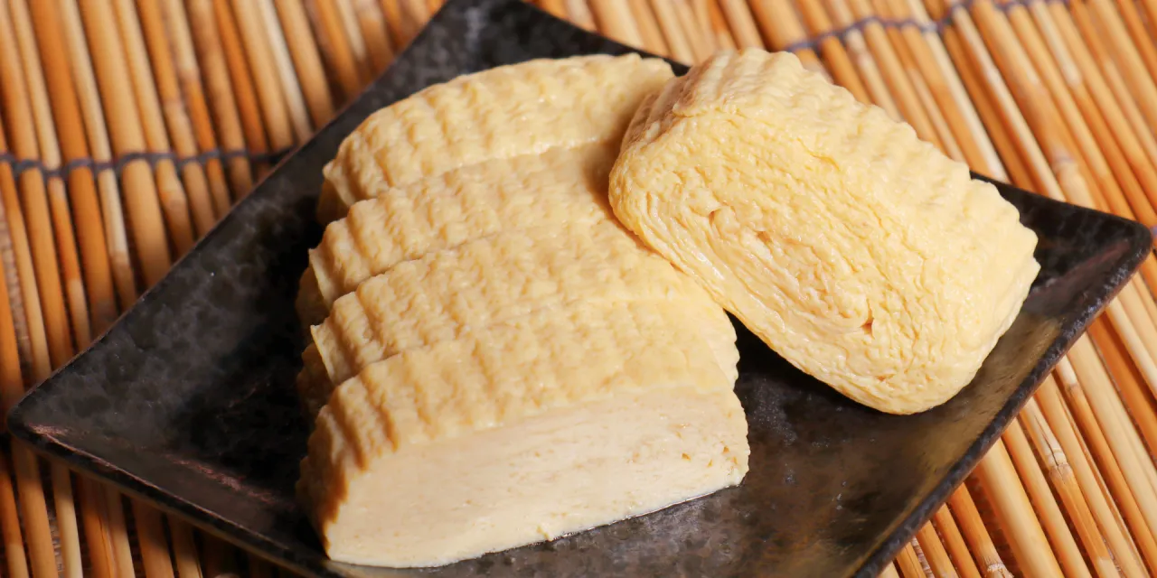 Na czarnym talerzu leżą pokrojone w plastry i jeden cały kawałek futomaki tamago, przygotowane z jajek, na tle bambusowej maty.