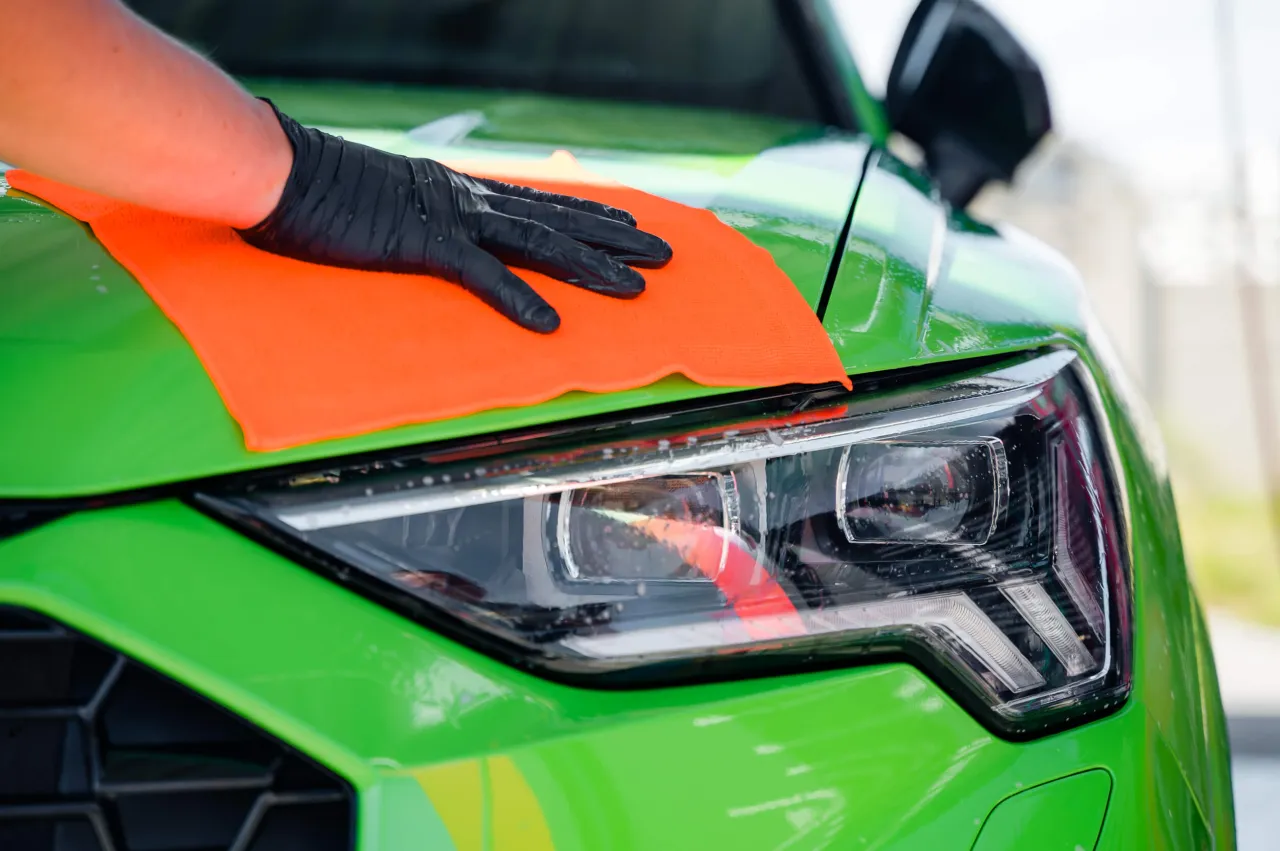 Ręka w czarnej rękawiczce z pomarańczową mikrofibrą wykonuje glinkowanie lakieru zielonego samochodu, usuwając zanieczyszczenia z reflektora.