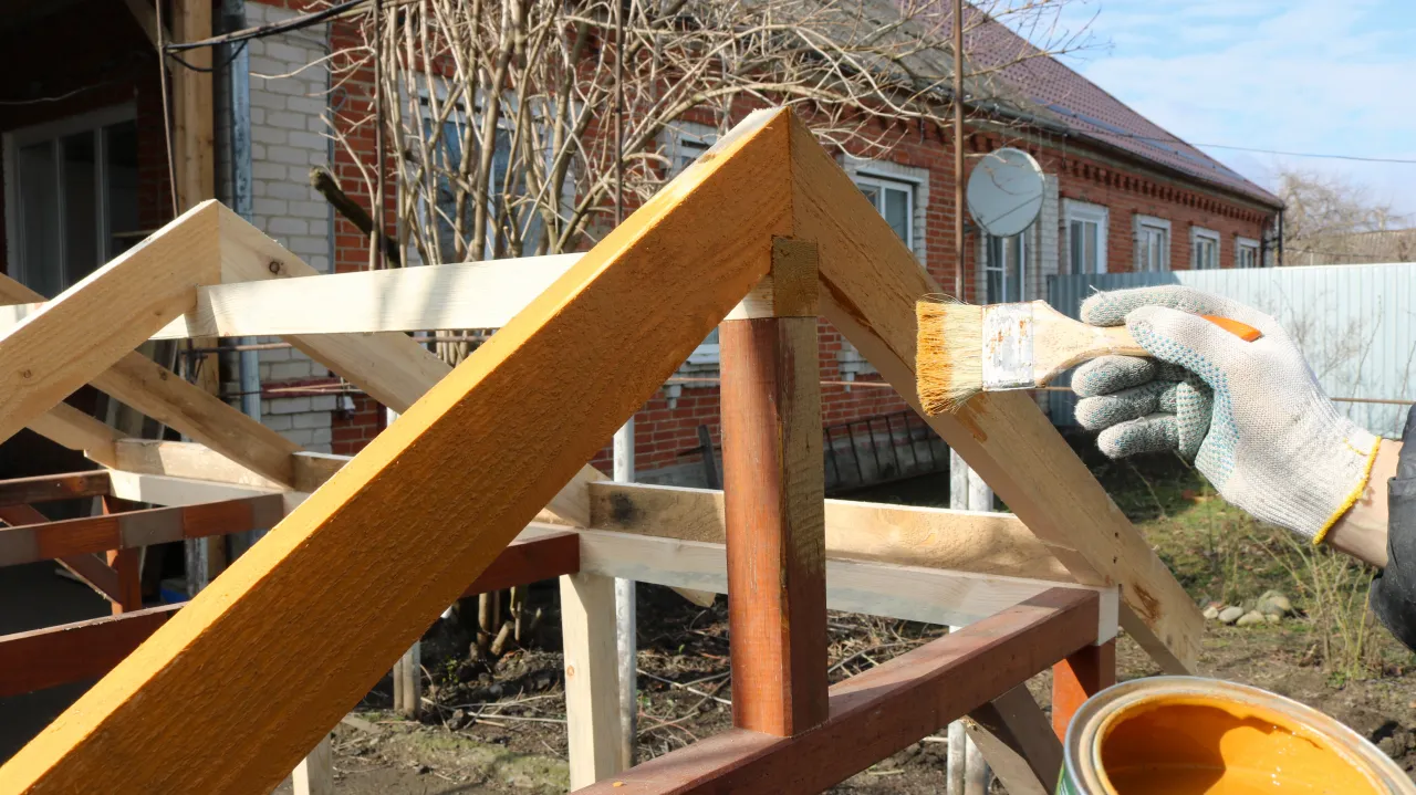 Malowanie drewnianej konstrukcji altany. Puszka z pomarańczową farbą i pędzel w rękawiczce. Idealne rozwiązanie, czym przykryć altanę, by ją zabezpieczyć.