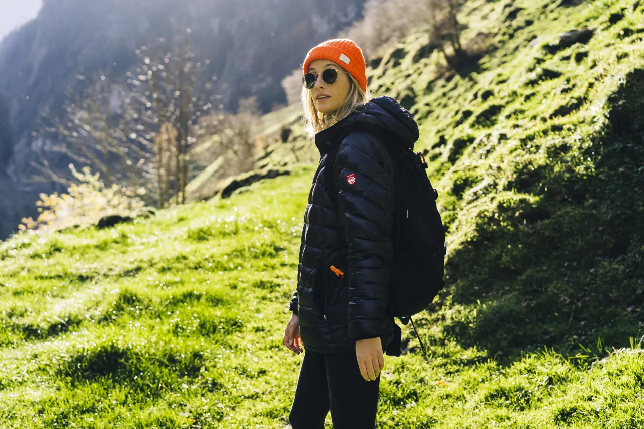 Kobieta w pomarańczowej czapce i czarnej kurtce z plecakiem na tle g&oacute;rskiego krajobrazu. Idealny zestaw, by wiedzieć, co spakować w g&oacute;ry.