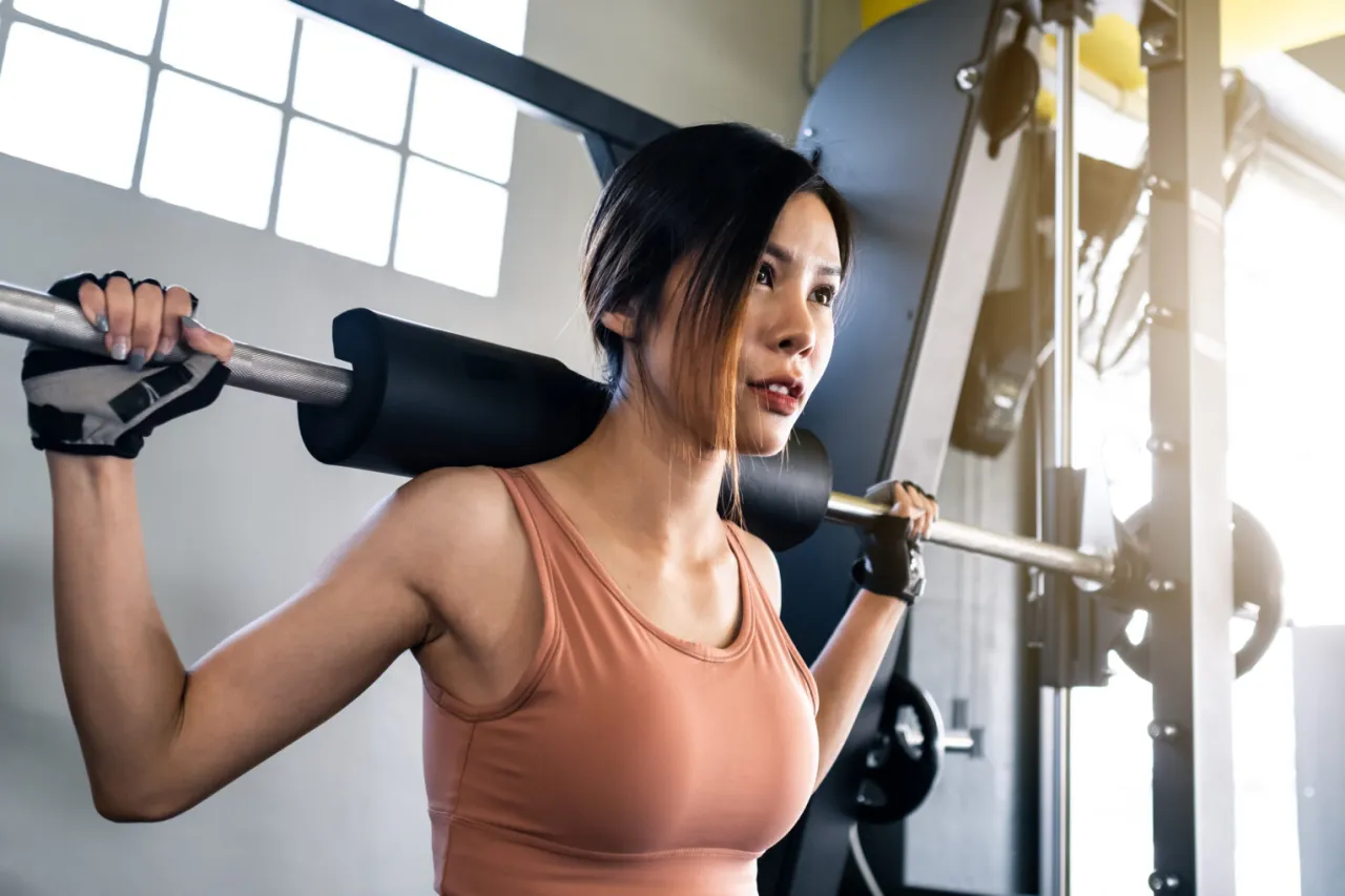 Kobieta z gryfem na barkach w siłowni. Pokazuje, jak połączyć crossfit z siłownią, budując siłę i wytrzymałość.