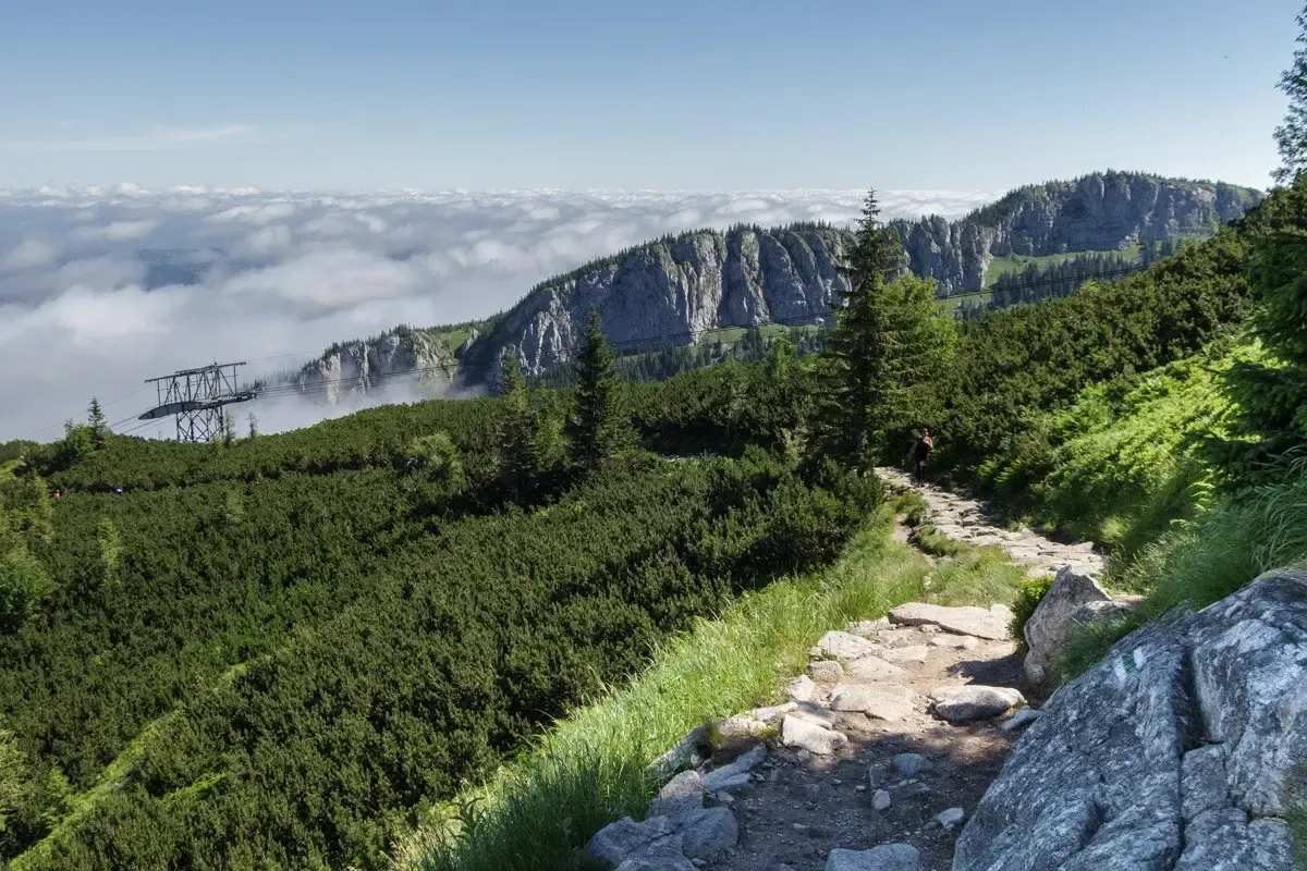 Kamienista ścieżka prowadzi przez zielone zbocza ku skalistym szczytom. W oddali widać morze chmur i konstrukcję kolejki na Kasprowy Wierch.