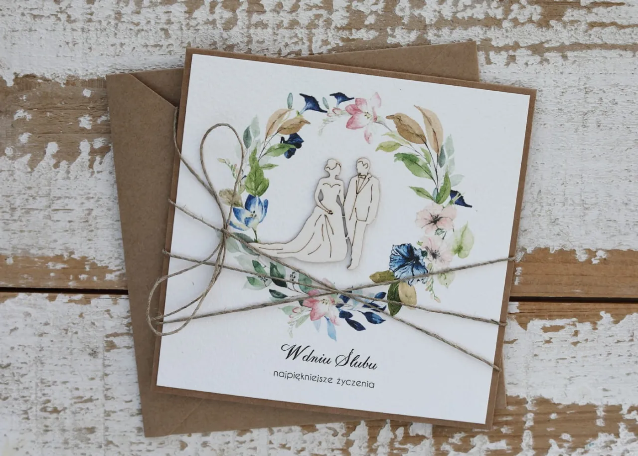 Elegancka kartka z życzeniami na ślub, ozdobiona wianuszkiem z kwiatów i sylwetkami pary młodej.