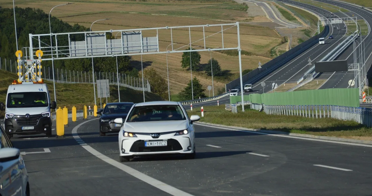 Biały samoch&oacute;d jedzie drogą ekspresową, kt&oacute;ra jest częścią trasy Krak&oacute;w-Miech&oacute;w. W tle widać inne pojazdy i krajobraz.