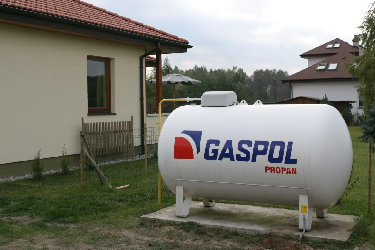 Zbiornik z napisem GASPOL PROPAN, kt&oacute;ry jest odpowiedzią na pytanie: jaki gaz do ogrzewania domu. W tle dom i drzewa.