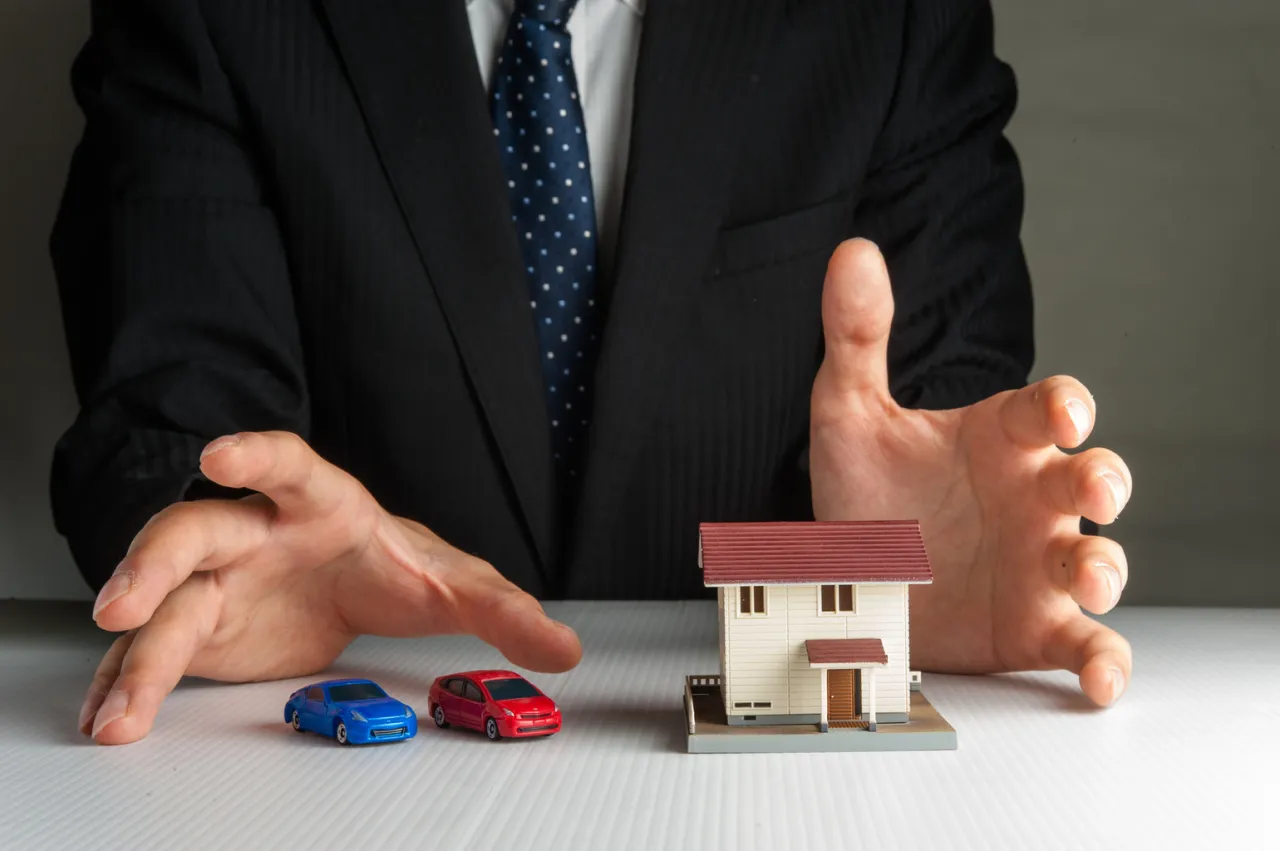 Ubezpieczenie domu a śmierć właściciela: dłoń chroni model domu i samochod&oacute;w, symbolizując bezpieczeństwo majątku po śmierci.