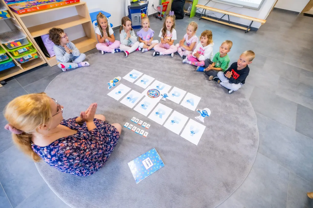 Nauczycielka z grupą dzieci na dywanie uczy się liter. Niska liczba dzieci na jednego opiekuna pozwala na indywidualne podejście.