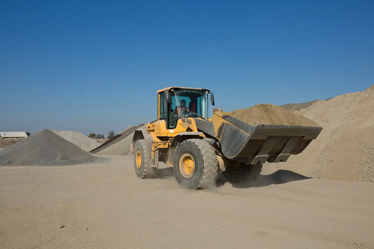 Żółta ładowarka Volvo z pełnym czerpakiem ziemi, jakby na egzamin na ładowarkę pytania i odpowiedzi. W tle hałdy kruszywa.