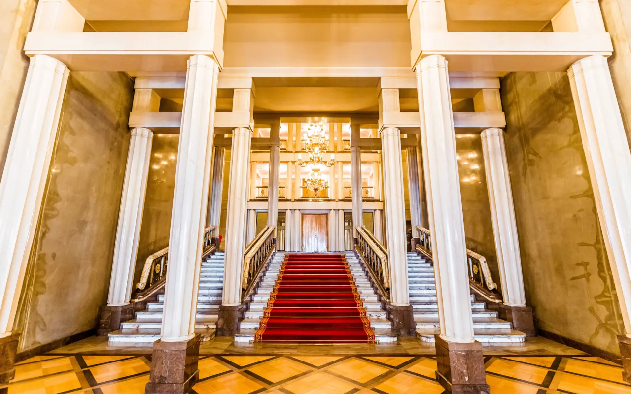 Eleganckie foyer teatru z czerwoną wykładziną na schodach, marmurowymi kolumnami i kryształowym żyrandolem.