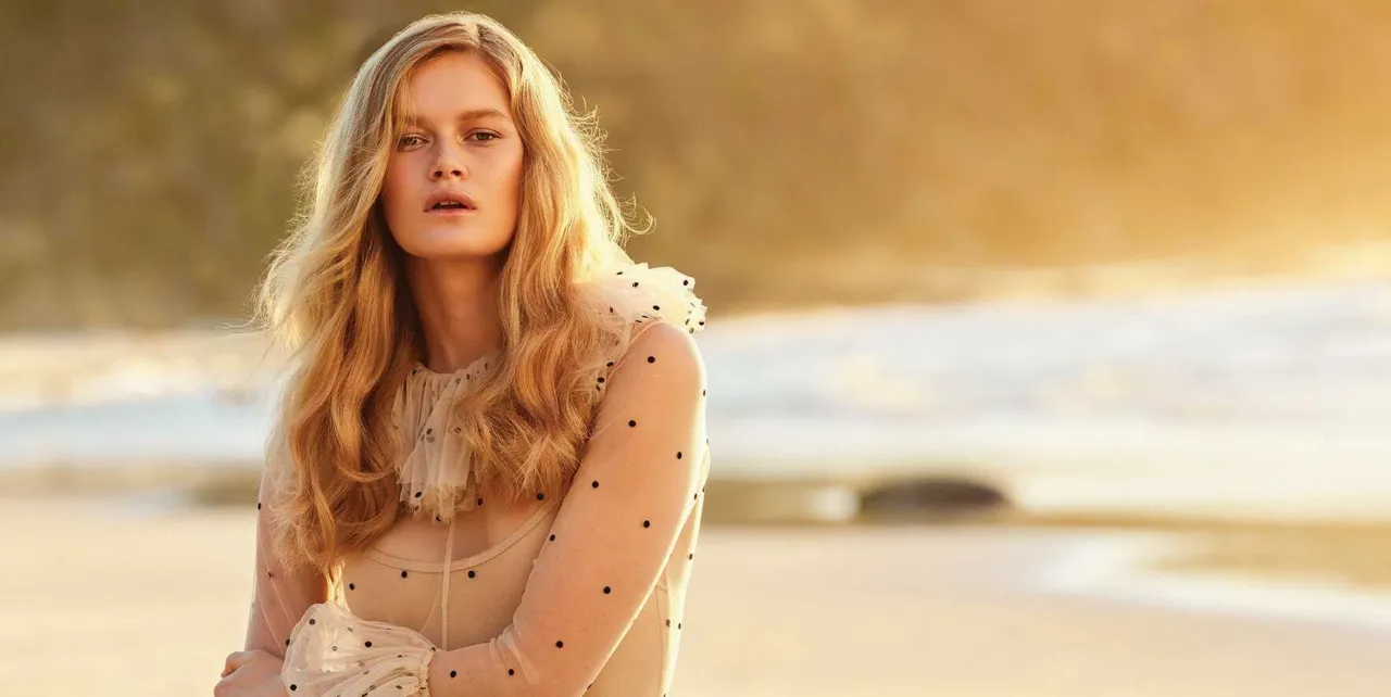 Kobieta z piękną, falowaną fryzurą średniej długości, w beżowej sukience w kropki, na plaży o zachodzie słońca.