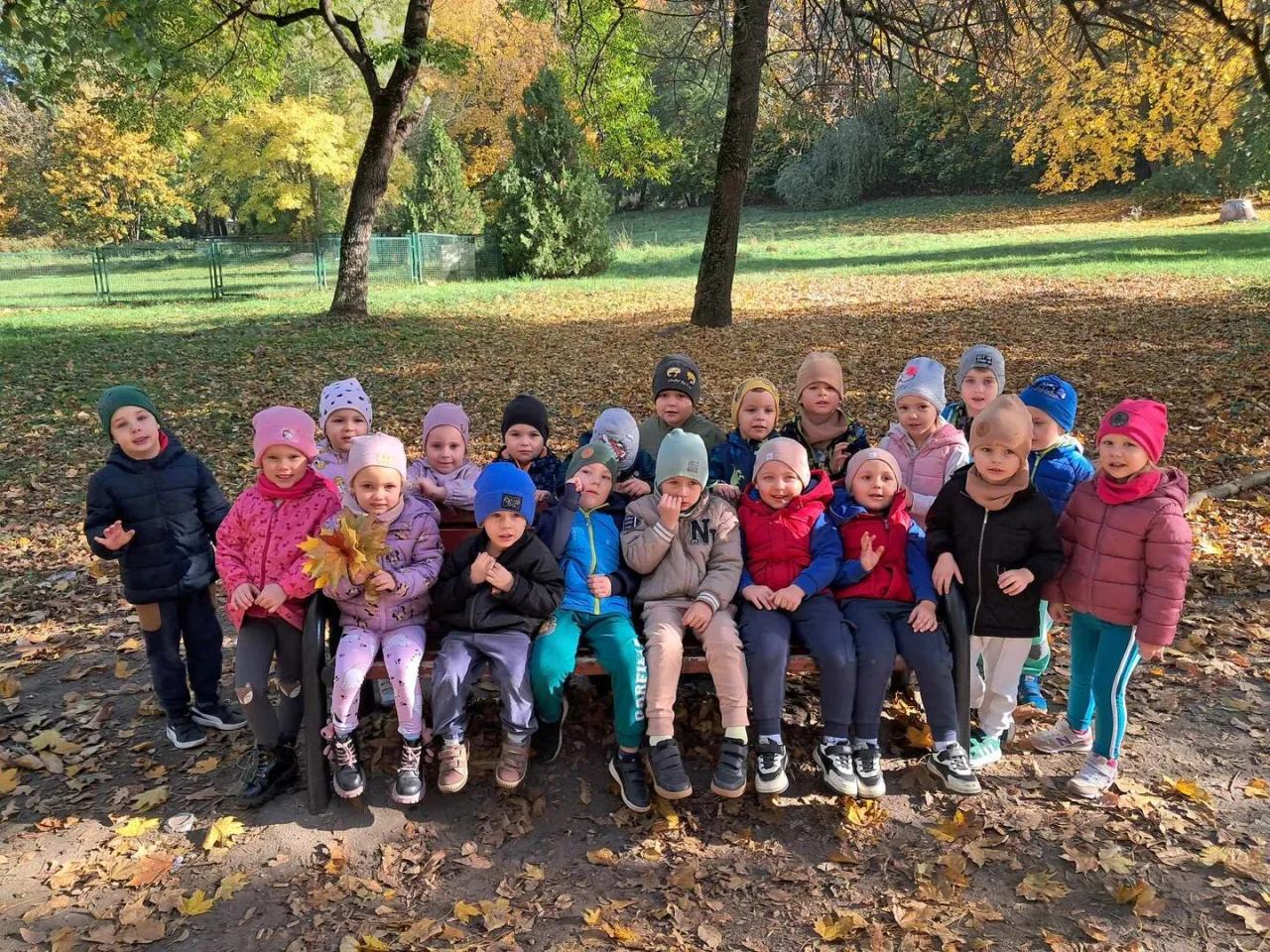 Dzieci w jesiennym parku, jak w wierszu o końcu lata. Liście sypią się z drzew, a maluchy pozują do zdjęcia.