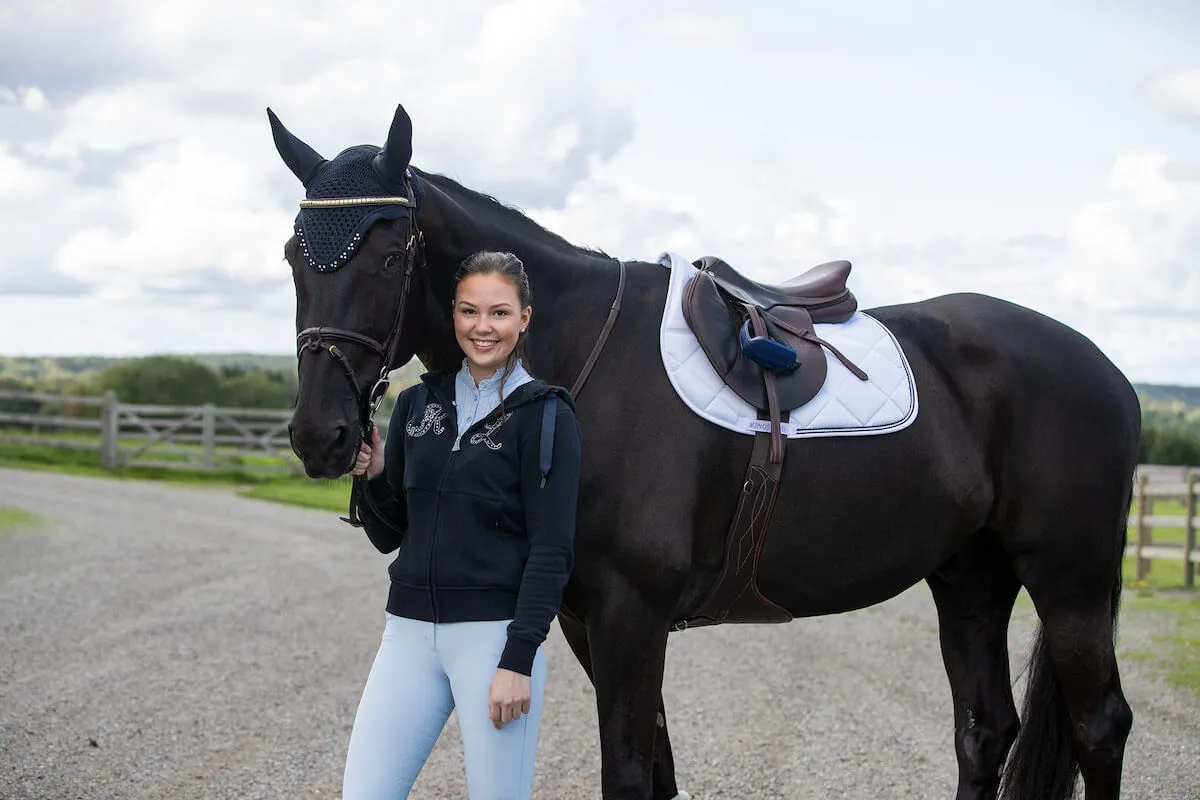 Uśmiechnięta dziewczyna stoi obok czarnego konia. Na grzbiecie konia widoczny jest biały czaprak z brązową siodłem.