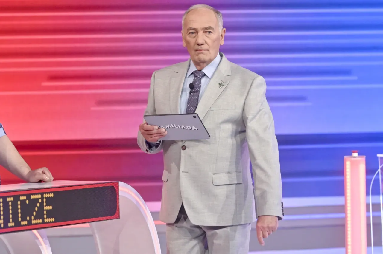 Prowadzący Familiady, ubrany w jasny garnitur, trzyma tablet z napisem 