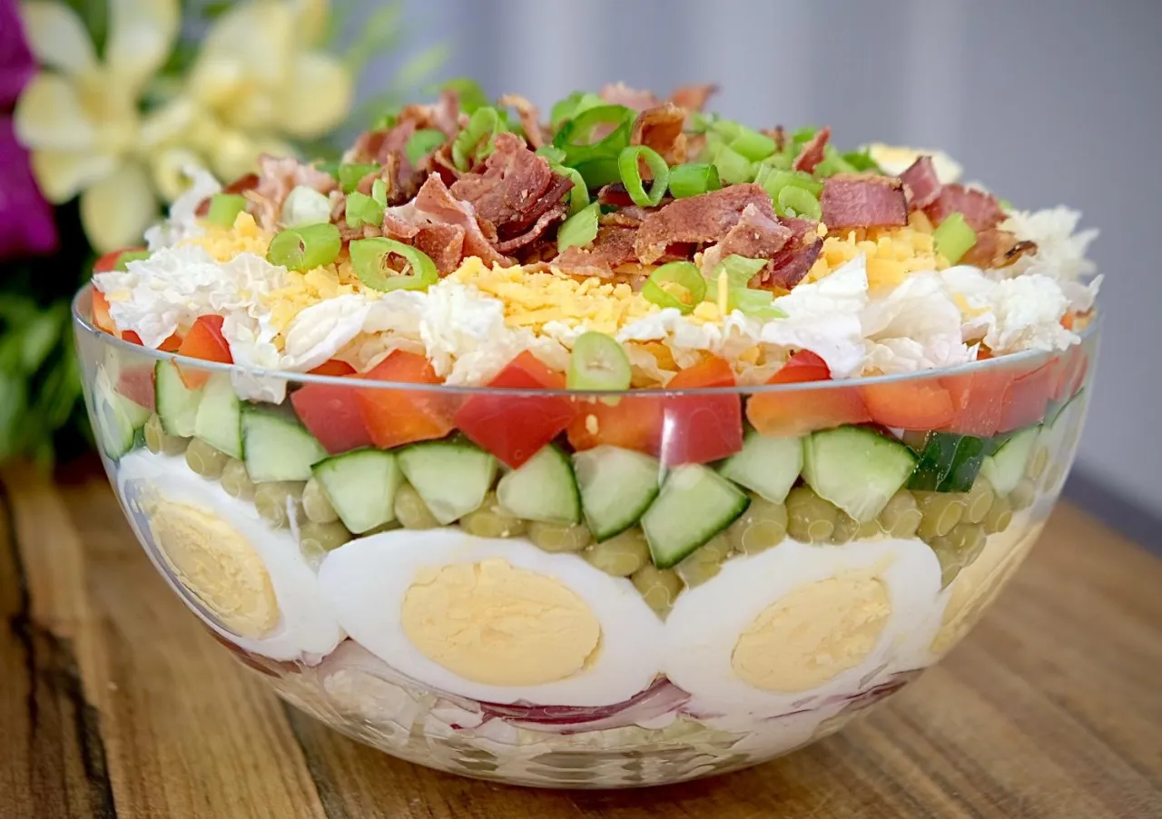 Warstwowa sałatka na Wielkanoc w szklanej misie: jajka, groszek, ogórek, pomidor, kapusta, ser, boczek i szczypiorek.