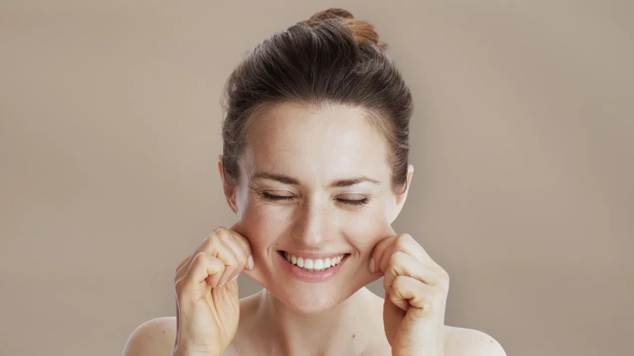 Uśmiechnięta kobieta po 40. z zamkniętymi oczami, delikatnie naciąga sk&oacute;rę na policzkach, pokazując jak wyglądać młodo.