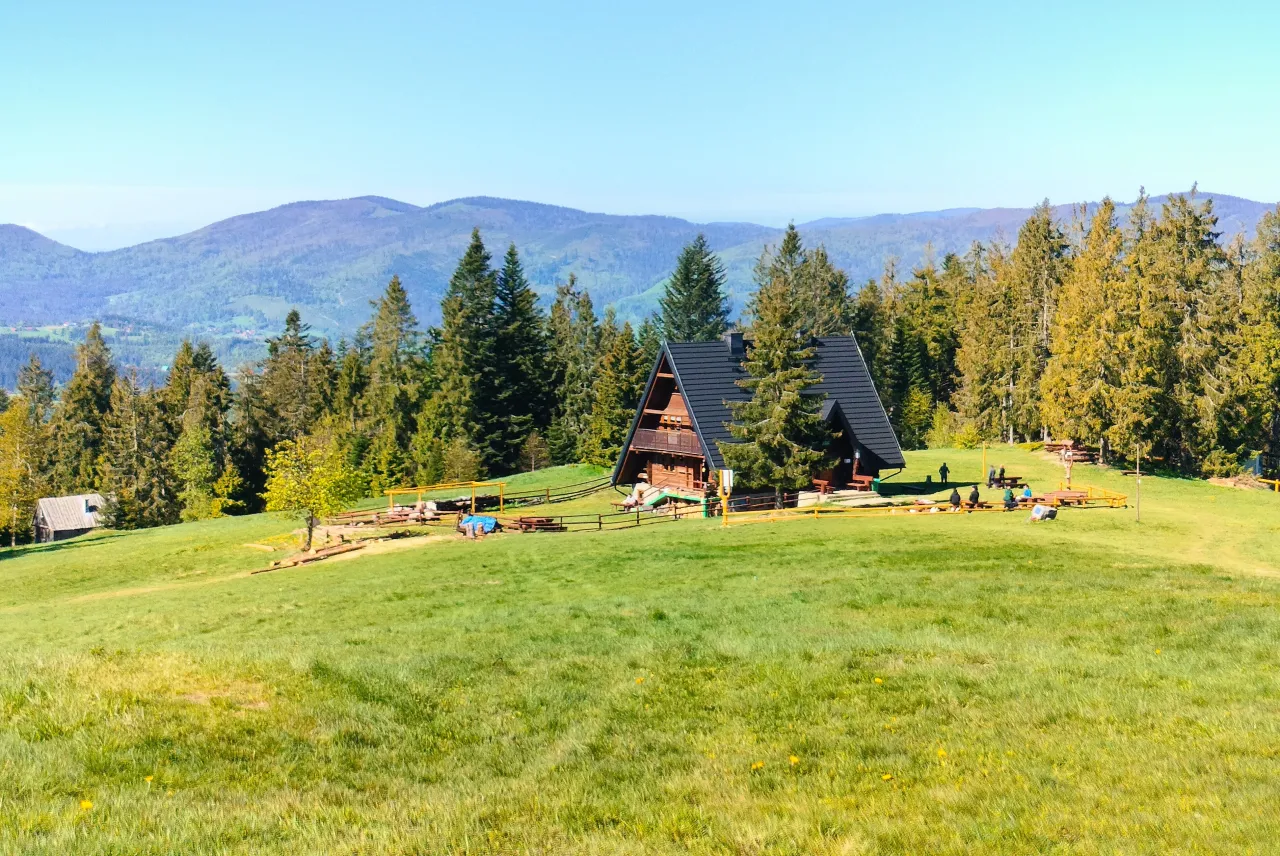 Malownicza bac&oacute;wka Krawc&oacute;w Wierch otoczona lasem i zielonymi łąkami, z widokiem na g&oacute;ry.