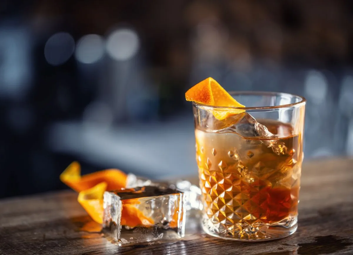 Gotowy drink Old Fashioned w szklance typu rocks z dużą kostką lodu i sk&oacute;rką pomarańczy