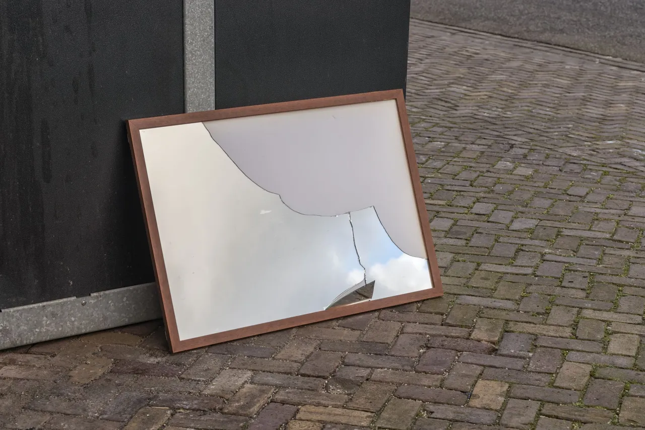 Rozbite lustro w drewnianej ramie leży przy czarnej ścianie. Zastanawiasz się, gdzie wyrzucić lustro?