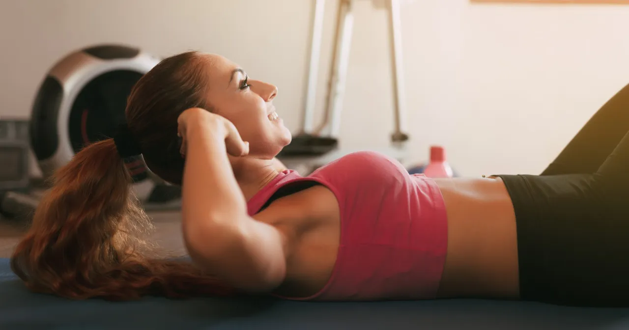 Uśmiechnięta kobieta wykonuje ćwiczenia na brzuch. W tle widać sprzęt do ćwiczeń. Czy codzienny trening ma sens? Tak, dla niej na pewno!