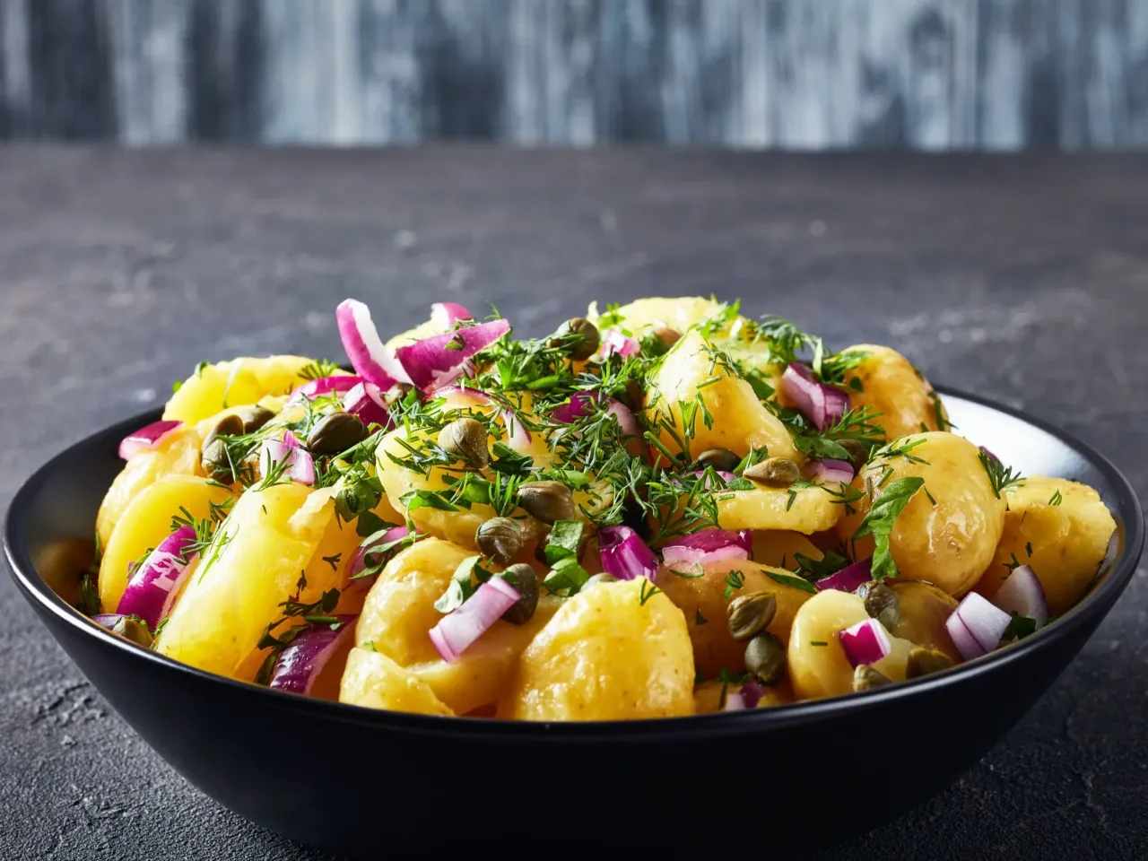 Sałatka ziemniaczana pysznie czy przepysznie? Ziemniaki, czerwona cebula, kapary i koperek w czarnej misce.