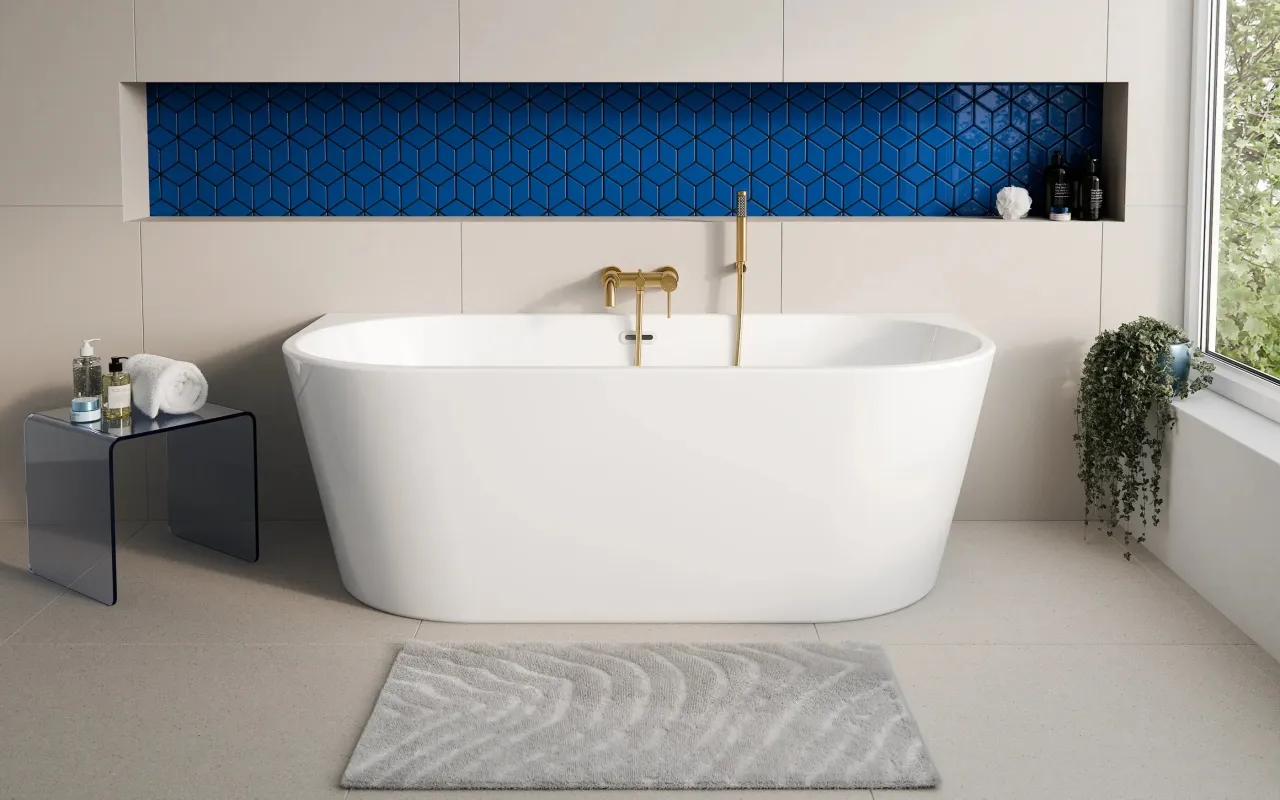 Wolnostojąca wanna z baterią w kolorze złota. Obok niej szklany stolik z kosmetykami i ręcznikiem. W tle niebieskie, geometryczne płytki. Zastanawiasz się, ile wanna ma litrów?