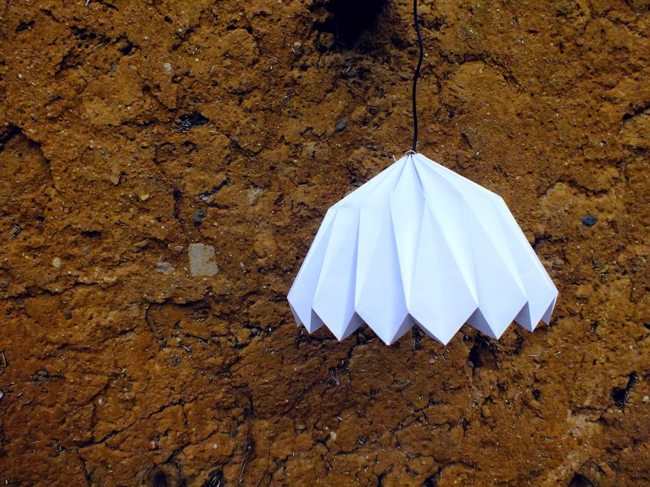 Biały abażur origami, kt&oacute;ry można zrobić samodzielnie, wisi na tle chropowatej, brązowej ściany.