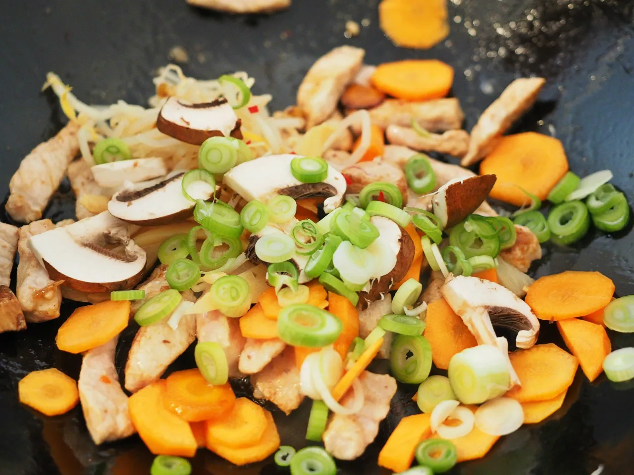 Pyszne dania z woka w przygotowaniu: kawałki kurczaka, pieczarki, marchewka i szczypiorek smażą się na patelni.