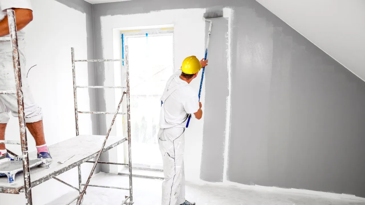 Malowanie sufitu na szaro. Pracownik w żółtym kasku maluje ścianę wałkiem. Obok rusztowanie.