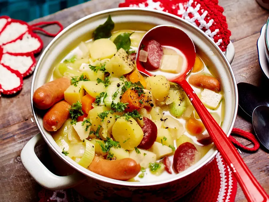 Großer Topf mit dampfender Kartoffelsuppe auf einem rustikalen Holztisch mit Löffeln und Schüsseln