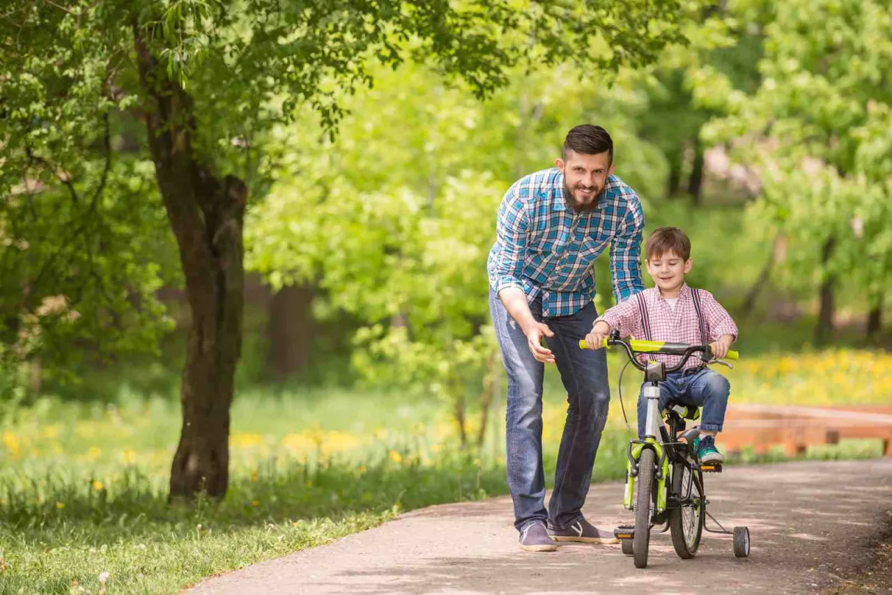 Ojciec uczy syna, który ma 10 latek, nie umie jeździć na rowerze. Chłopiec siedzi na rowerze z bocznymi kółkami, a tata go asekuruje.
