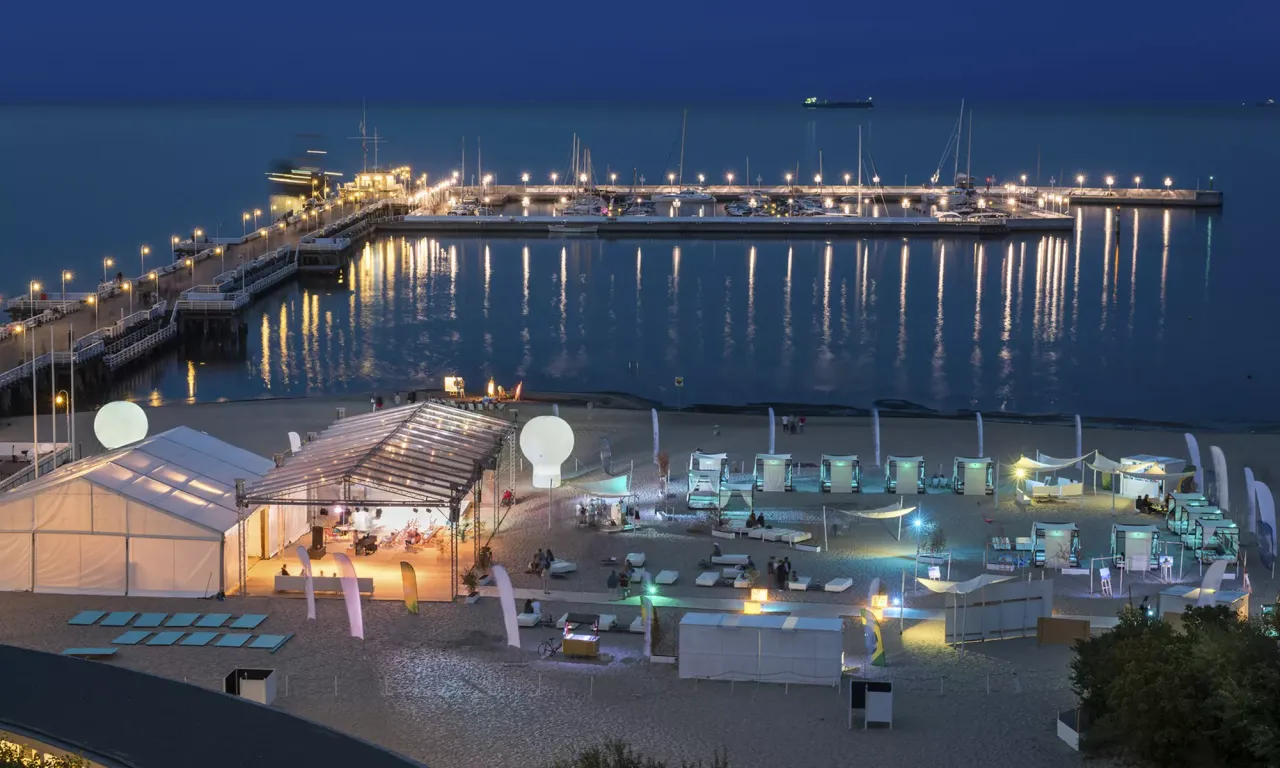 Wieczorny spacer po molo i plaży w Sopocie. Widać tam namioty, leżaki i łodzie w marinie.