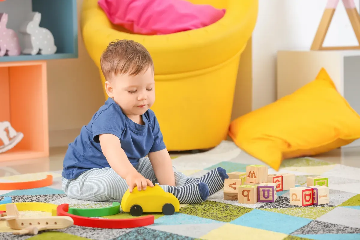 Małe dziecko bawi się ż&oacute;łtym autkiem i klockami na kolorowym dywanie. Myśli o tym, do kiedy można liczyć na dofinansowanie do żłobka.