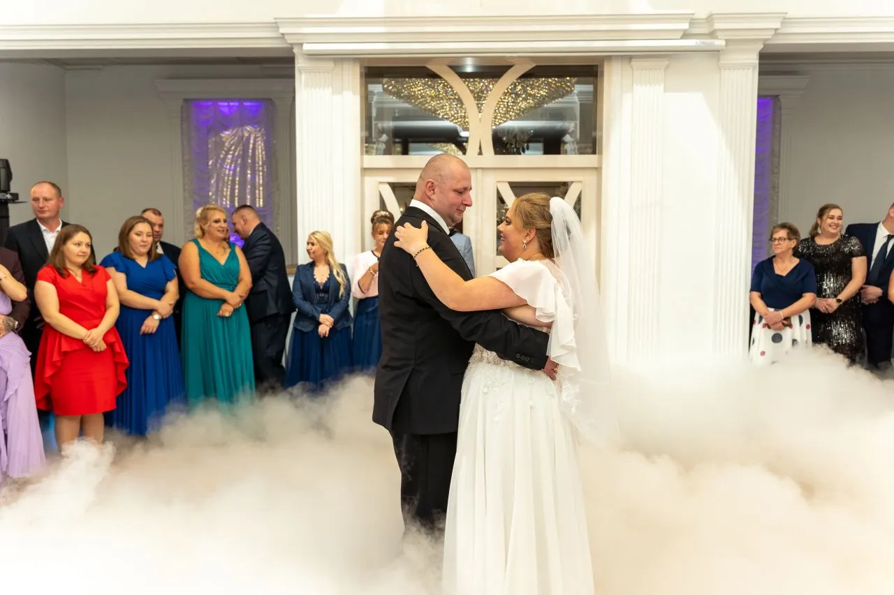Para imprezy na kt&oacute;re wynajmujemy fotografa: para młoda tańczy w chmurach dymu, goście weselni obserwują.