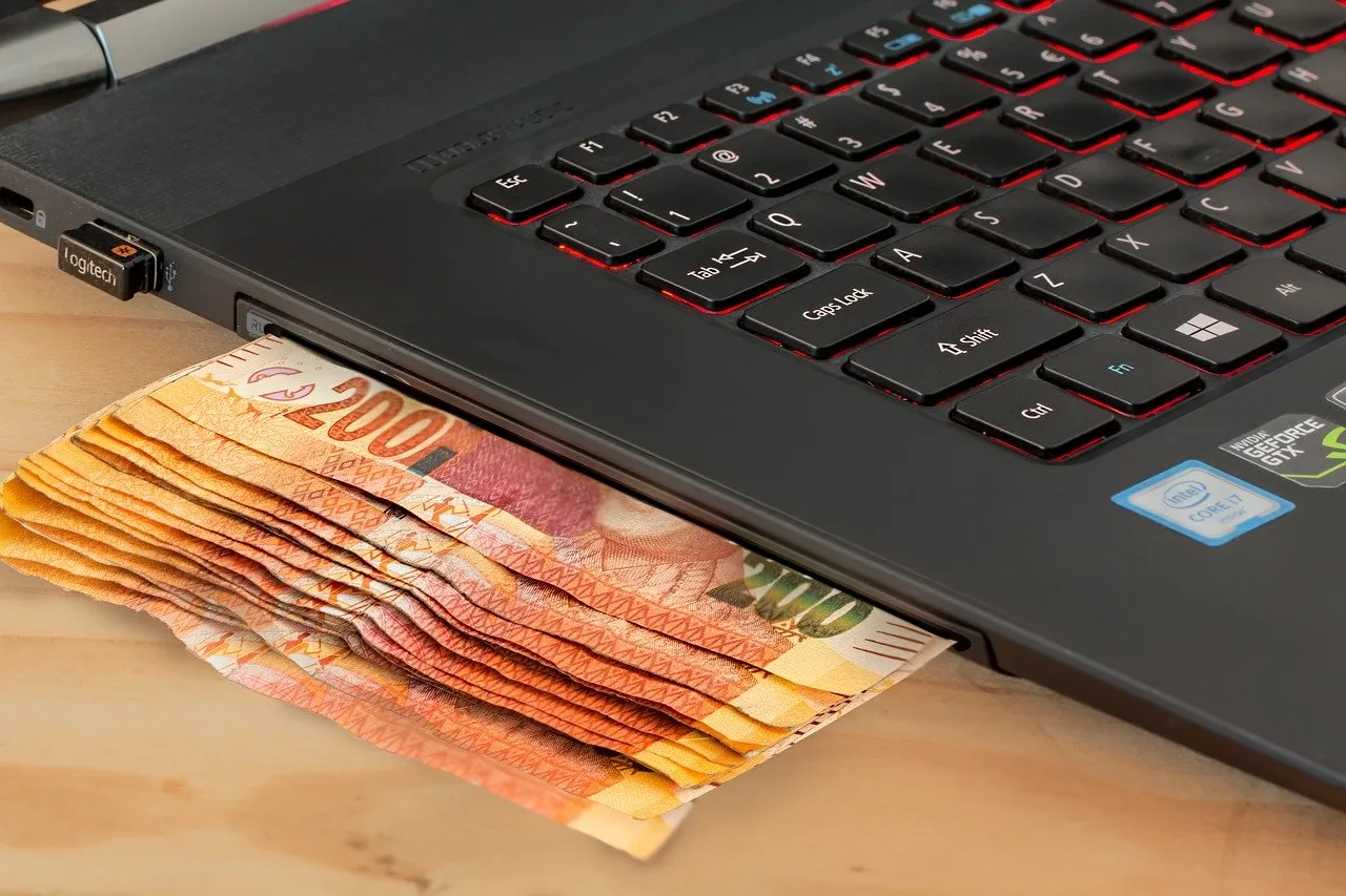 Stos banknot&oacute;w wsuwa się pod laptopa z podświetlaną klawiaturą. Zastanawiasz się, ile kosztuje laptop?