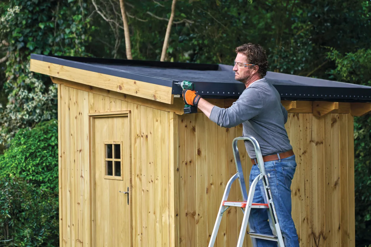 Mężczyzna na drabinie przykręca dach do domku ogrodowego. Pokazuje, jak zrobić dach w domku ogrodowym.
