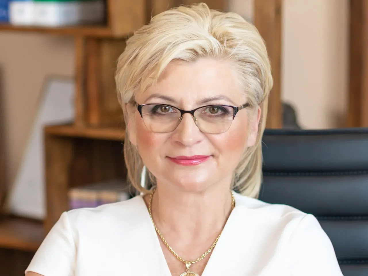 Kobieta w okularach, w białej bluzce i złotej biżuterii, siedzi w gabinecie. To może być przykład sukcesji w biznesie.