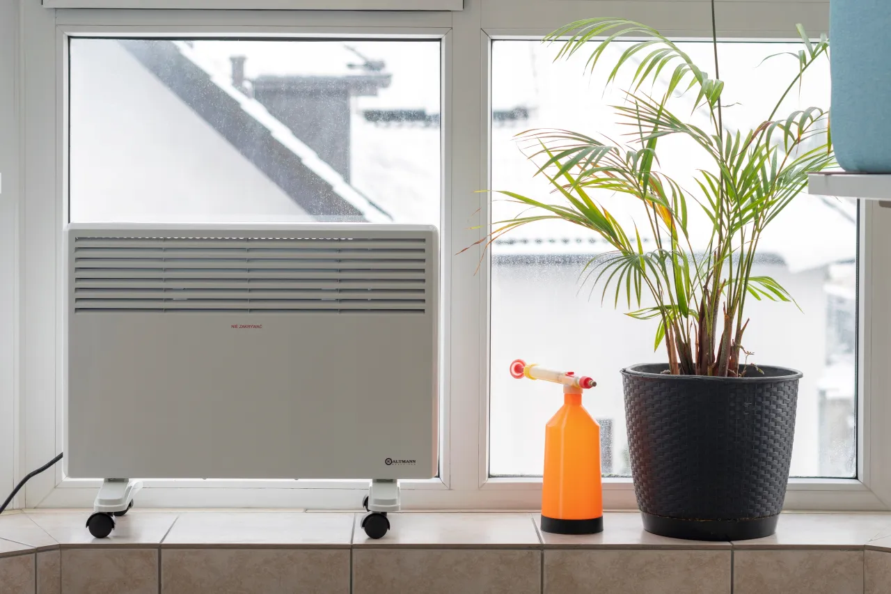 Jaki grzejnik do pokoju 15m2? Na parapecie stoi biały grzejnik na k&oacute;łkach, obok doniczka z palmą i pomarańczowy spryskiwacz.