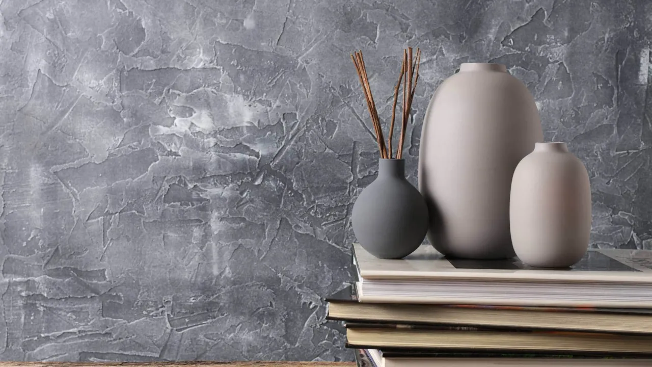 Szara ściana z efektem tynku dekoracyjnego, na której stoją książki z wazonami. Inspiracja, jak położyć tynk dekoracyjny.