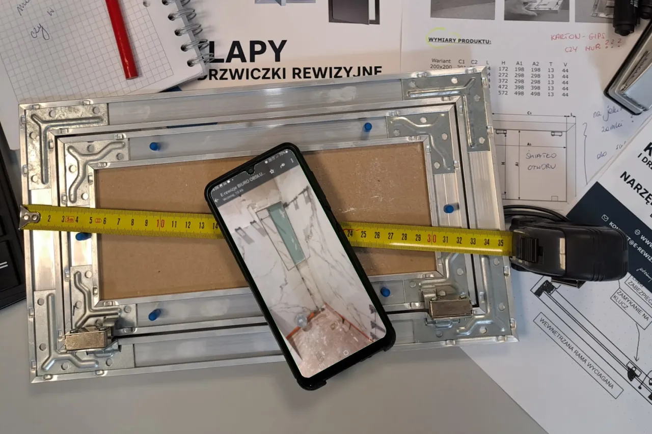 Instrukcja jak zamontować drzwiczki rewizyjne plastikowe. Miarka i telefon z podglądem pokazują wymiary.