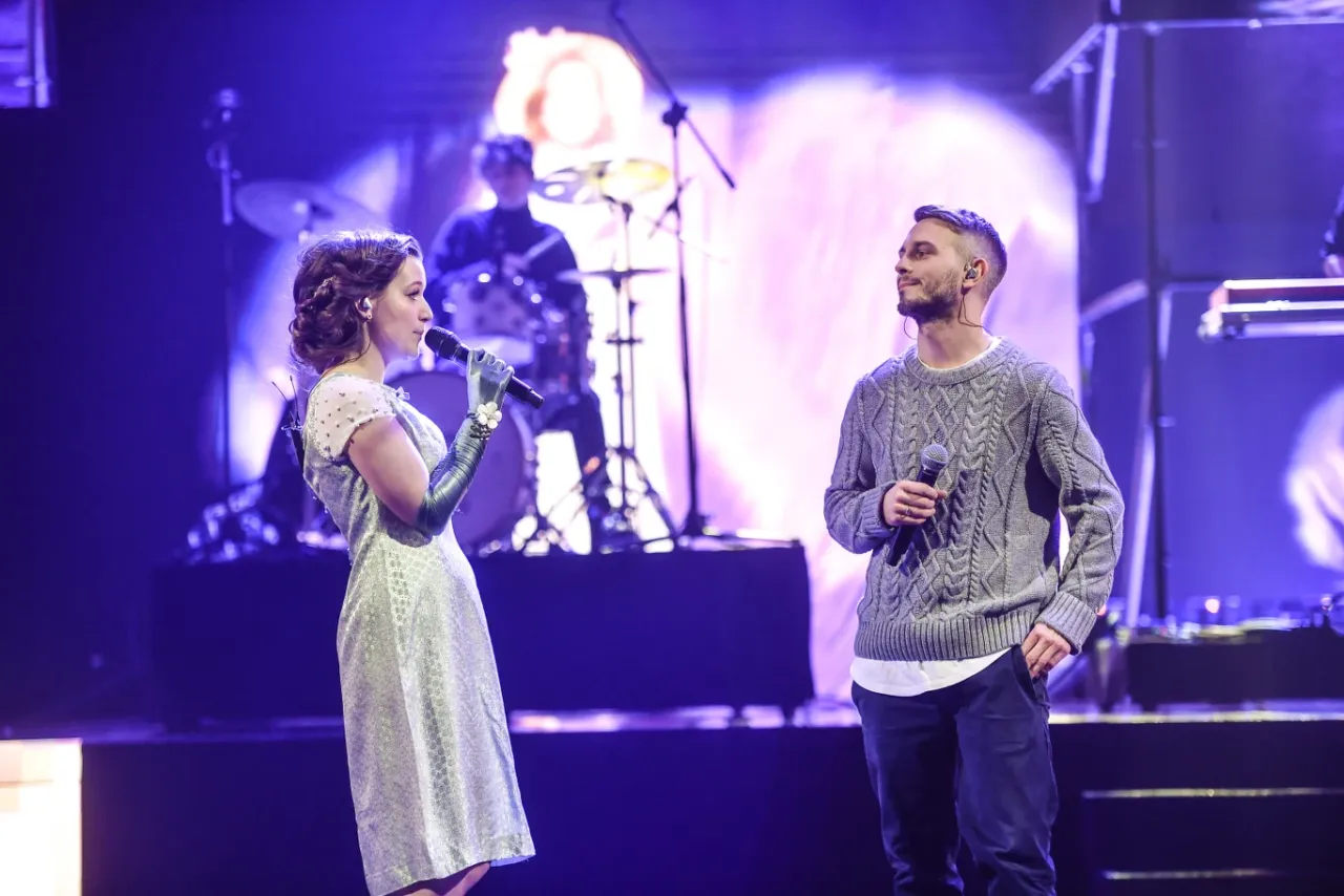 Kto śpiewa kawka na wynos? Wokalistka w srebrnej sukience i długich rękawiczkach oraz wokalista w szarym swetrze występują na scenie.