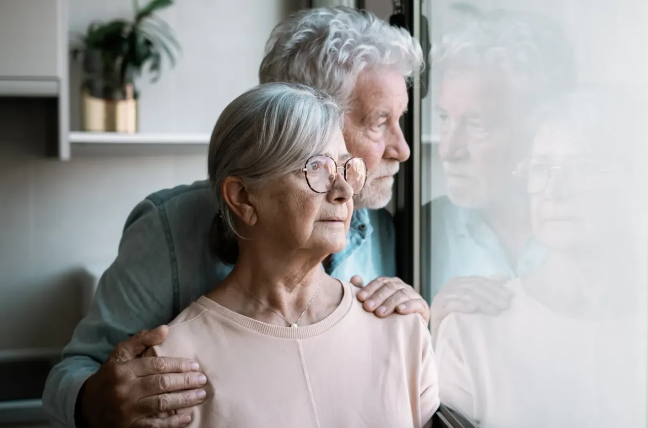 Para, kto to jest senior obecnie? Para starszych ludzi patrzy przez okno, mężczyzna obejmuje kobietę ramieniem.