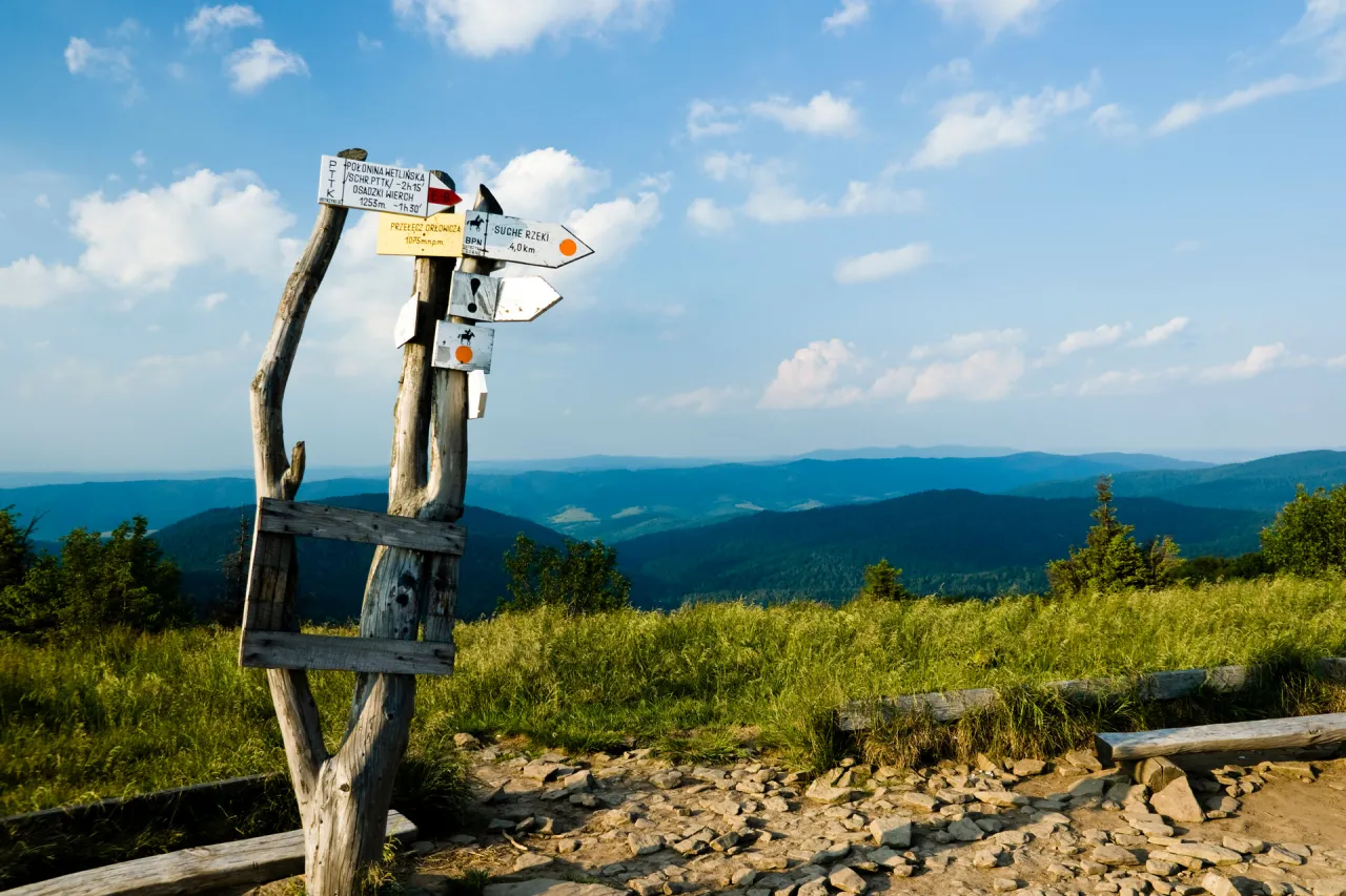 Gdzie w g&oacute;ry na maj&oacute;wkę? Znaki wskazują szlaki, a za nimi rozciągają się zielone wzg&oacute;rza i błękitne niebo.