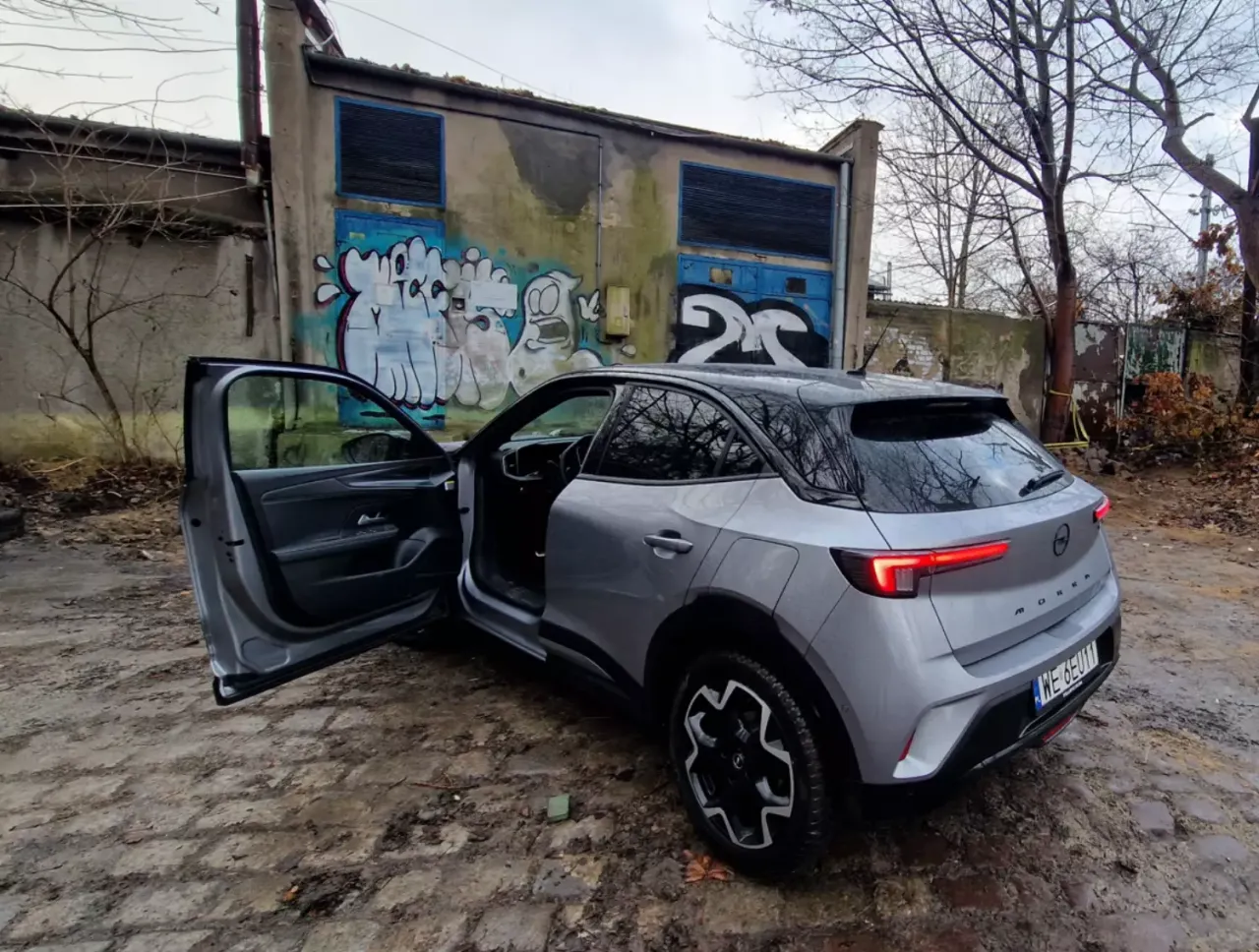 Srebrny Opel Mokka z otwartymi drzwiami, gotowy do wycofania z firmy i darowizny. Tło to zniszczony budynek z graffiti.